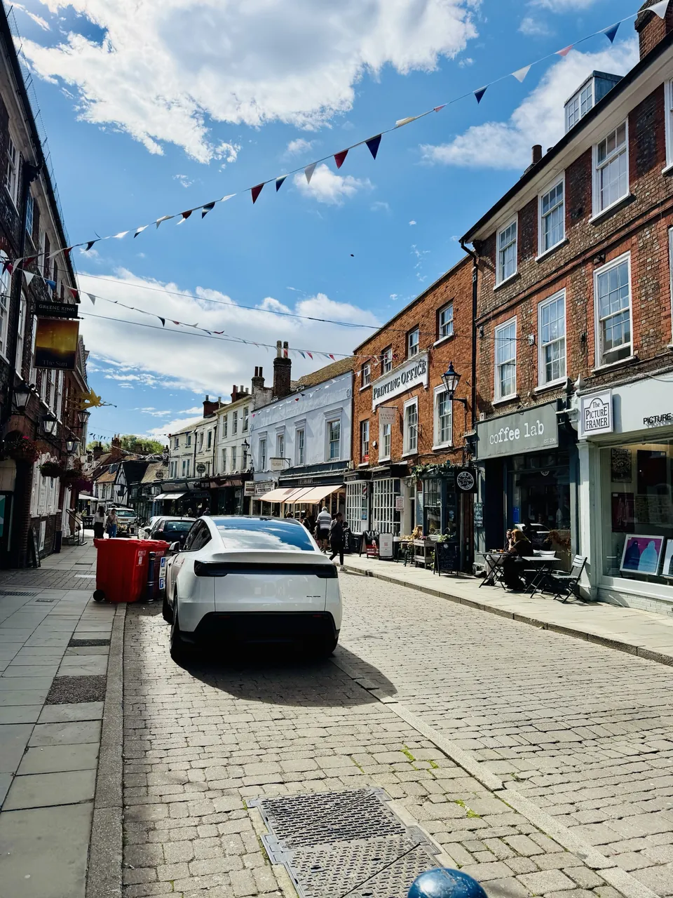 Bunting strung across a Hitchin street of brick and painted shopfronts, a coffee shop and a picture framer open, people at pavement tables.